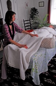 Client reclining on table with white blanket as healer runs energy through hands into clients body at ankles and knees, plants in background