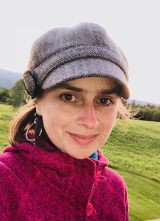 Head shot of woman with dark brown eyes, a gray hat and a fushia sweater with mountains in the background