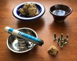 Moxa Pole on silver colored holder, loose moxa in blue and white dish, cup of water, stick on moxa and moxa cone on wooden table