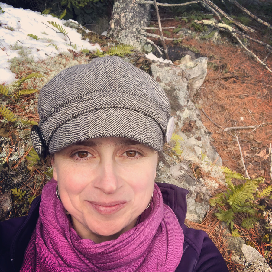 Headshot of woman smiling while wearing a gray hat with a brim, purple scarm and jacket, with a backdrop of a moss covered white quartz boulder and the forest floor