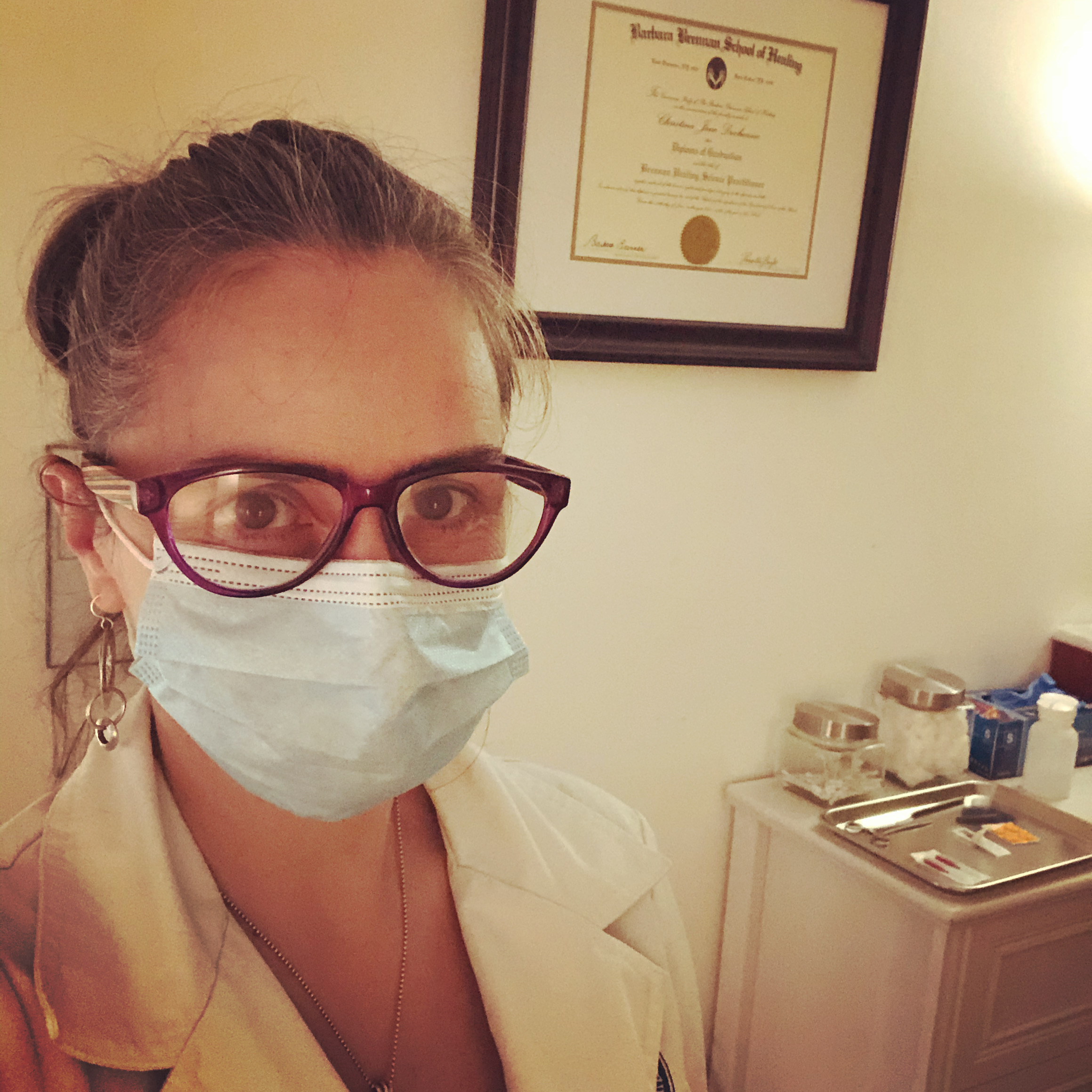 Woman with glasses and hair tied back wearing a mask and a lab coat in an acupuncture office