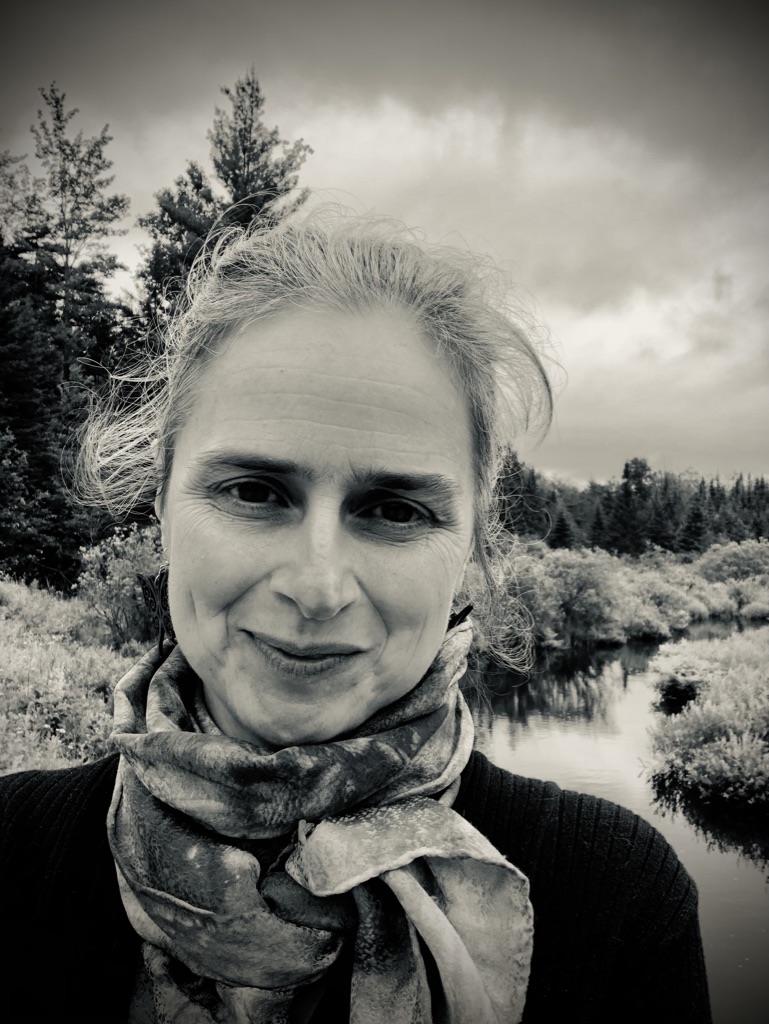 Black and white photo of woman in front of a winding stream with hair pulled back, smiling, and a scarf wound around her neck.